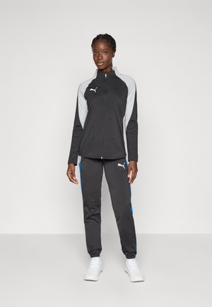 Black athletic jacket with grey accents, zip closure, and logo. Paired with black pants featuring blue side stripes. White sneakers.