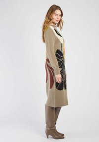 Robe pull en beige avec des motifs floraux audacieux en noir, jaune et rouge. Manches longues, poignets et ourlet côtelés, portée avec des bottes marron jusqu'aux genoux.