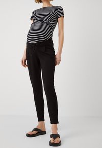 Graviditetskläder bestående av en svart och vit randig kortärmad topp samt svarta byxor med dragsko, tillsammans med svarta flip-flops.