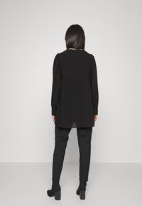 Tunic nera con tessuto texturizzato, maniche lunghe e retro plissettato. Abbinata a pantaloni neri a sigaretta e stivaletti con tacco chunky.