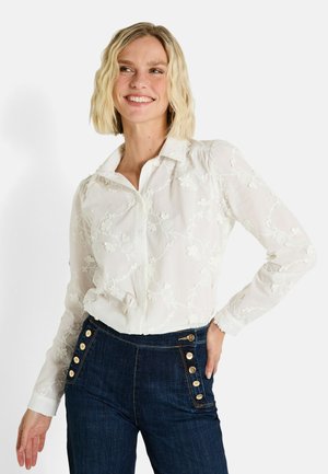 Blouse blanche brodée avec des motifs floraux, à manches longues et col. Associée à un pantalon en denim foncé taille haute avec des boutons dorés.