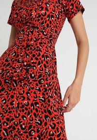 Czerwona sukienka w panterkę z krótkimi rękawami, podkreśloną talią i gładką teksturą materiału. Posiada czarne i jasnoróżowe wzory.