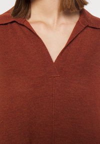 Poitrine et cou d'une femme portant un pull en tricot couleur rouille à col en V avec un détail simple au niveau du col.