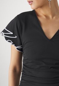 Musta V-purkka T-paita lyhyillä röyhelöhihoilla, jossa on valkoista brodeerausta. Pehmeä kangas ja poimuttaminen sivuilla.