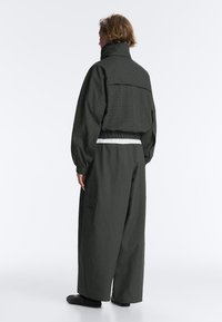 Personne aux cheveux courts portant une veste ample texturée sombre et un pantalon large, debout de profil sur un fond blanc.