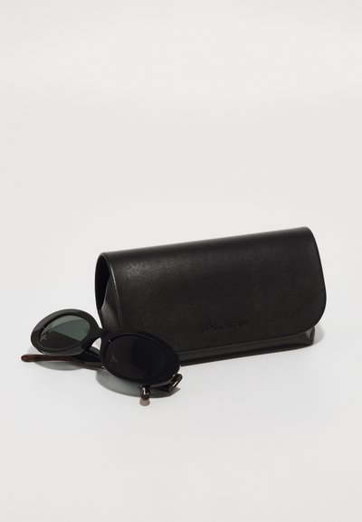 Schwarze Sonnenbrille mit runden Gläsern und dicken Rahmen neben einem mattschwarzen Lederetui mit Klappenverschluss und eing prägendem Branding.