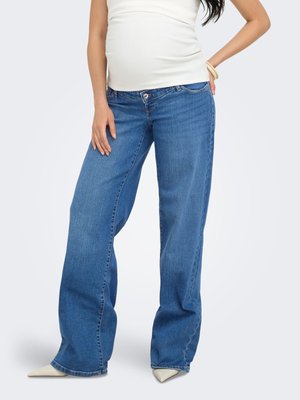 Femme enceinte portant un jean bleu à jambes larges, un haut blanc, des chaussures blanches à bout pointu et un bracelet, debout avec une main sur la hanche.