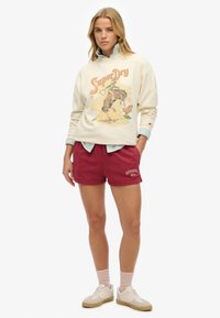 Felpa crema con grafica "SuperDry" e design colorato. Pantaloni corti bordeaux con logo. Sneakers beige chiaro e calzini rosa completano l'outfit.