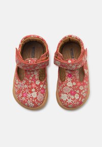Červené dětské boty s květinovým vzorem, se suchým zipem, zaoblenou špičkou a hnědou gumovou podrážkou. Obsahují malé bílé a růžové květinové vzory.