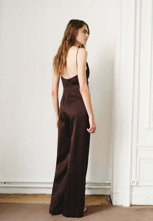 Femme aux longs cheveux ondulés portant une combinaison sans manches et dos nu de couleur marron foncé, debout à l'intérieur contre un mur blanc et un encadrement de porte.