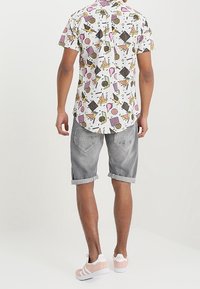 Hombre que lleva una camisa blanca de manga corta con patrones geométricos coloridos, pantalones cortos de mezclilla gris remangados y zapatillas rosas y blancas.