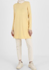 Nainen, joka pitää yllään vaaleankeltaista pitkähiharaista tunikaa, kermaisia housuja, beigejä tennareita ja valkoista huivia yksinkertaista taustaa vasten.