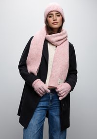 Rosa ribbad stickad mössa, scarf och handskar i kombination med en krämfärgad kabelstickad tröja och svart jacka; blå denimjeans med en etikettdekoration.