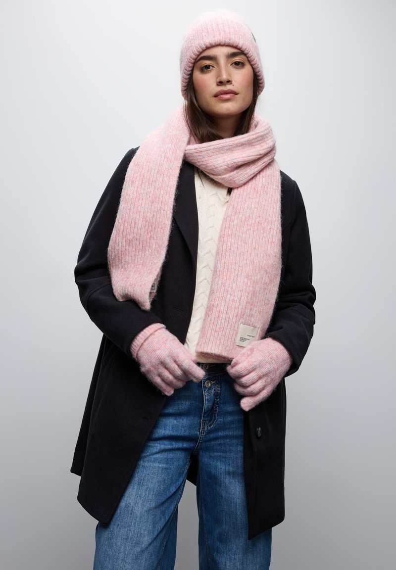 Rosa ribbad stickad mössa, scarf och handskar i kombination med en krämfärgad kabelstickad tröja och svart jacka; blå denimjeans med en etikettdekoration.