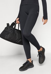 Μαύρα εφαρμοστά κολάν από ελαστικό ύφανση, συνδυασμένα με μαύρο ζιβάγκο που κλείνει με φερμουάρ και ελαφριά μαύρα αθλητικά παπούτσια. Κρατάει μια μαύρη τσάντα γυμναστηρίου.