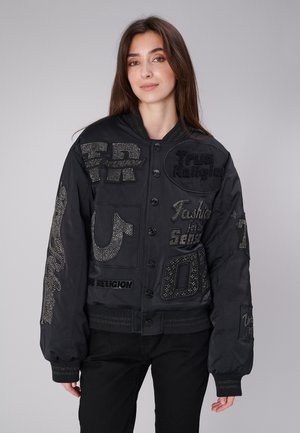 Jeune femme portant une veste noire boutonnée avec des patchs de texte ornés et un pantalon noir, debout devant un fond gris uni.