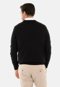 Maglione nero con scollatura rotonda e polsini a costine, indossato sopra una camicia bianca con colletto. Abbinato a pantaloni beige chiaro.