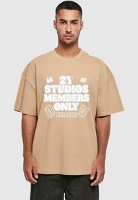 Beige oversize t-shirt med korte ærmer og tekst på forsiden: "2Y STUDIOS MEMBERS ONLY EST. 2010." Materialet ser blødt ud, og designet er afslappet.