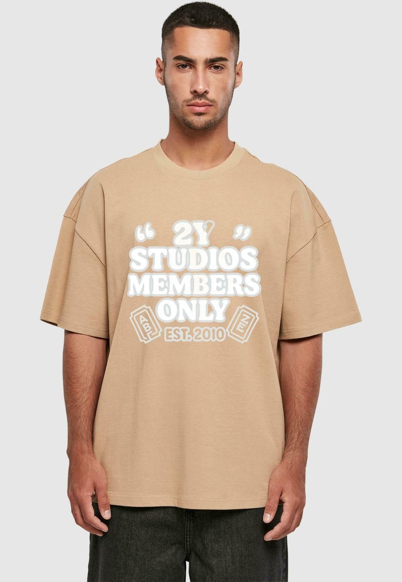 Beige oversized tričko s krátkým rukávem a nápisem vpředu: "2Y STUDIOS MEMBERS ONLY EST. 2010." Materiál vypadá měkce, design je neformální.