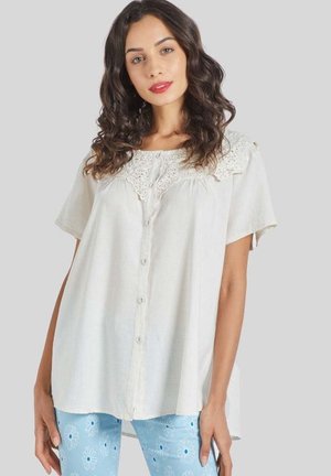 Mujer con cabello rizado oscuro, vistiendo un top blanco con botones y detalles de encaje, y pantalones azul claro con patrones florales blancos sobre un fondo gris.