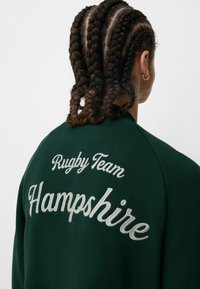 Sudadera de color verde oscuro con texto bordado que dice "Equipo de Rugby Hampshire", con costuras redondeadas en los hombros y una textura de tela suave.