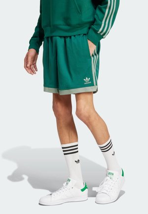 Osoba nosząca zielone krótkie spodenki i bluzę Adidas, białe skarpetki Adidas z czarnymi paskami oraz białe buty sportowe z zielonymi detalami.
