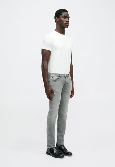 Γκρι στενά τζιν και λευκή κοντομάνικη μπλούζα, συμπληρωμένα με μαύρα δερμάτινα παπούτσια. Τα τζιν έχουν ήπια φθορά και πέντε τσέπες.