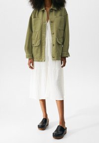 Olivově zelená plátěná bunda s velkými kapsami, nošená přes strukturované bílé šaty. Černé dřevěné klíny dokreslují celý outfit.
