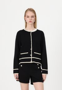 Black cardigan with cream trim, featuring a round neckline, front buttons, and two pockets. Paired with black shorts.