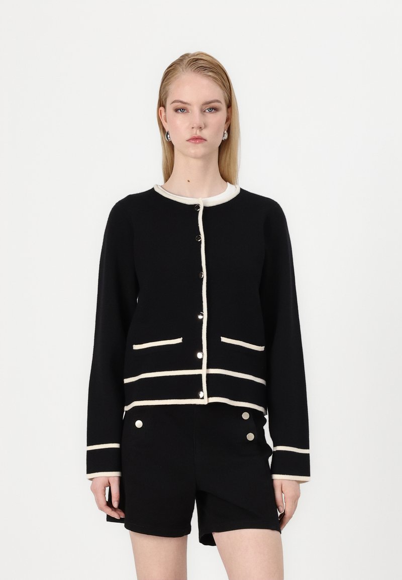 Black cardigan with cream trim, featuring a round neckline, front buttons, and two pockets. Paired with black shorts.