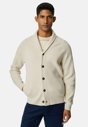 Cardigan beige a costine con colletto a scialle, bottoni neri e tasche laterali, abbinato a una camicia a righe leggera sotto.