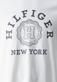 Tricou din bumbac alb, cu un logo negru ce include textul „HILFIGER” și „NEW YORK”, având un emblema circulară și detalii decorative.