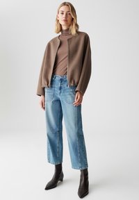 Frau, die eine braune Zip-Jacke über einem braunen Rollkragenpullover, hellblauen hochgeschnittenen Jeans und schwarzen spitzen Knöchelschuhen trägt, vor einem weißen Hintergrund steht.