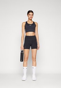 Juodas sportinis liemenėlis ir šortai, pagaminti iš tamprių medžiagų. Modelis laiko juodą vandens butelį. Baltas Nike logotipas ant liemenėlio, šortų ir kojinių.