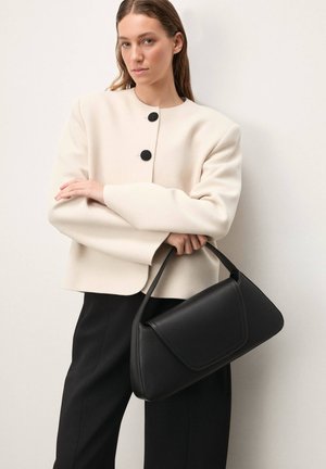 Black leather handbag with a curved shape and flap closure, held by a person wearing an ivory, round-neck jacket with black buttons.