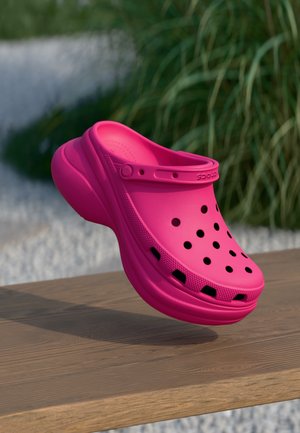 Leuchtend pinker Clog zum Hineinschlüpfen mit Lüftungslöchern und Riemen, schwebend über einer Holzoberfläche mit verschwommenem grünem Laubhintergrund.