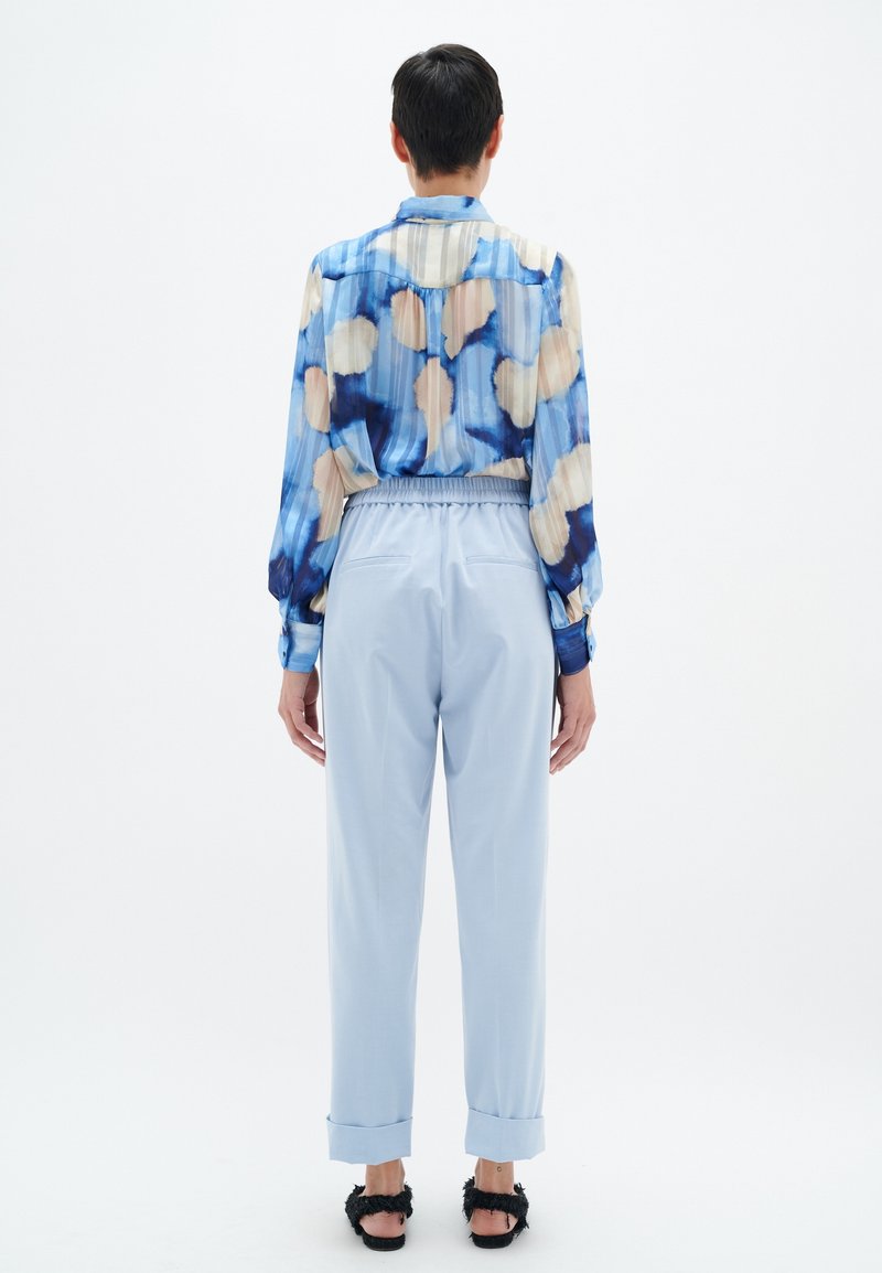 Personne aux cheveux courts portant un chemisier à motifs bleu et beige, un pantalon taille haute bleu clair et des sandales noires duveteuses, vue de dos.