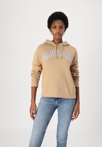 Haut à capuche en coton beige avec cordon de serrage, décoré d'une broderie "GANT 1949" en bleu. Assorti à un jean en denim bleu. Texture lisse et coupe décontractée.
