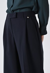 Navy-Hosen mit gefaltetem Design, taillierter Taille und glatter Textur. Goldfarbener Akzentknopf am Bund sichtbar.