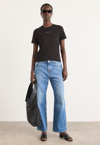 Svart kortermet t-skjorte med logo, lyseblå vide jeans, svart tote bag og svarte sandaler, i en nøytral bakgrunn.