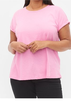 Femme portant un t-shirt à manches courtes rose clair et un pantalon noir, les mains doucement jointes devant elle.