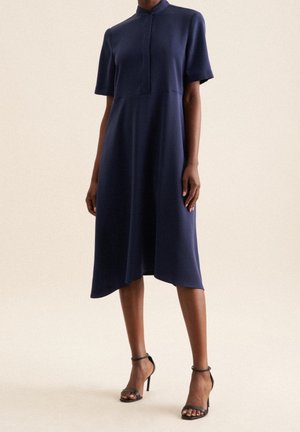 Frau trägt ein marineblaues, kurzärmeliges, knielanges Kleid mit asymmetrischem Saum und schwarzen High-Heel-Sandalen, steht vor neutralem Hintergrund.