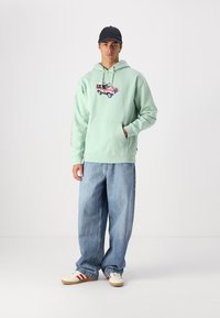 Světle zelená mikina s grafickým potiskem auta, přední kapsou a stahovací kapucí. V kombinaci s volnými světle modrými džíny a bílými teniskami.