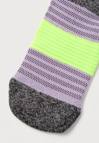 Zeķes pirksta zona tumši pelēkā krāsā ar rakstainu tekstūru, blakus violetām un melnām horizontālām svītrām, virs kurām atrodas spilgta neona zaļa josla.