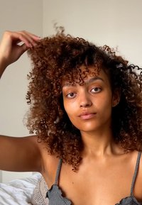 Kiharat ruskeat hiukset, joissa tiukat ja selkeät kiharat, vaalea ihonväri ja minimaalinen meikki. Päällä harmaa teksturoitu toppi, jossa säädettävät olkaimet.