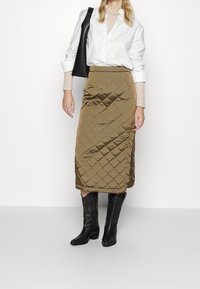 Gepatchter olivgrüner Rock mit einem seitlichen Schlitz, kombiniert mit einem weißen Hemd mit Knöpfen und schwarzen Cowboystiefeln. Strukturierter Stoff, knielanges Design.