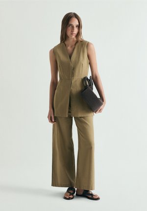Femeie care poartă o vestă olive fără mâneci și pantaloni largi, ținând o geantă de mână texturată închisă la culoare, stând pe un fundal simplu și luminos.
