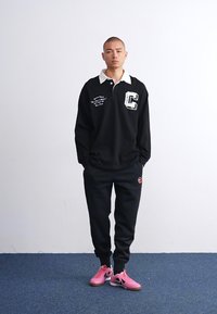 Svart langermet polo shirt med hvit krage og "C"-logo, kombinert med svarte joggebukser og rosa treningssko. Enkelt design, uformell passform.