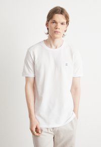 Tricou alb din bumbac, cu decolteu rotund, mâneci scurte și un mic detaliu brodat "II" de culoare albastră pe partea stângă a pieptului. Textură netedă.