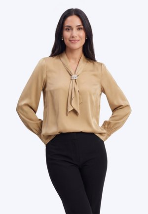 Frau mit langen dunklen Haaren, die eine satinierte beige Bluse mit Perlenbrosche und schwarze Hose trägt, steht mit den Händen in der Hüfte und lächelt.